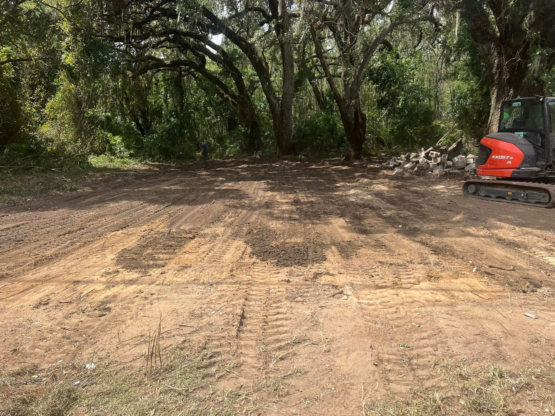 An orange excavator sits on a freshly cleared patch of dirt in front of a wooded area.
