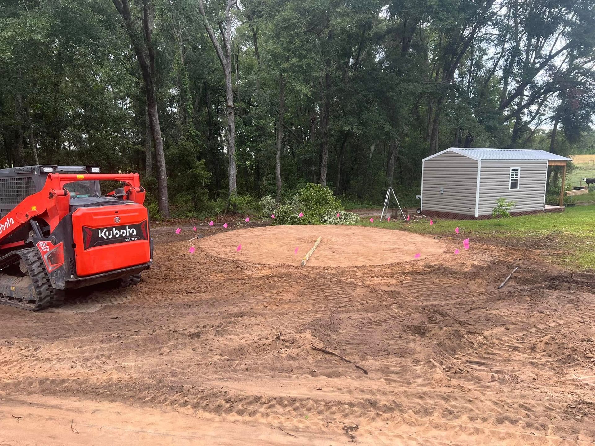 An orange skid steer parked beside a freshly leveled, circular dirt pad marked with pink flags in a rural yard.