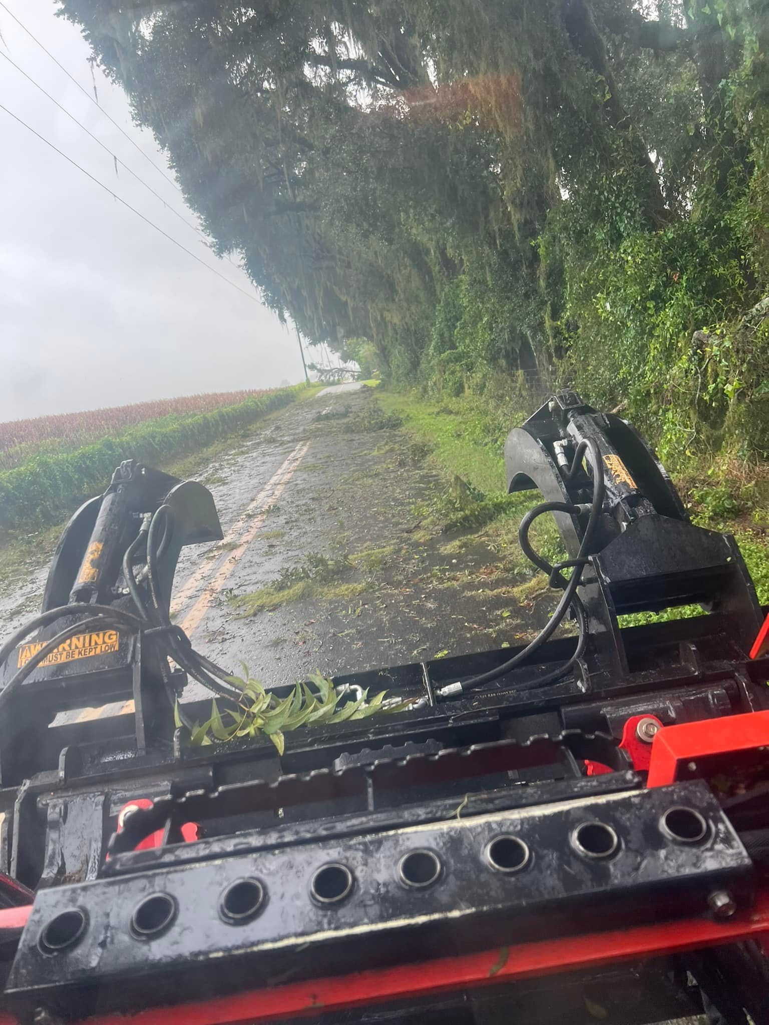 A first-person view from a tractor clearing a narrow, rain-slicked country road bordered by a dense tree line and a field.