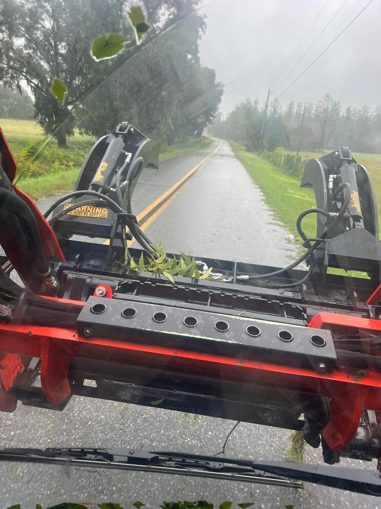 First-person view from inside a machine with red mechanical arms clearing branches from a wet, tree-lined road.