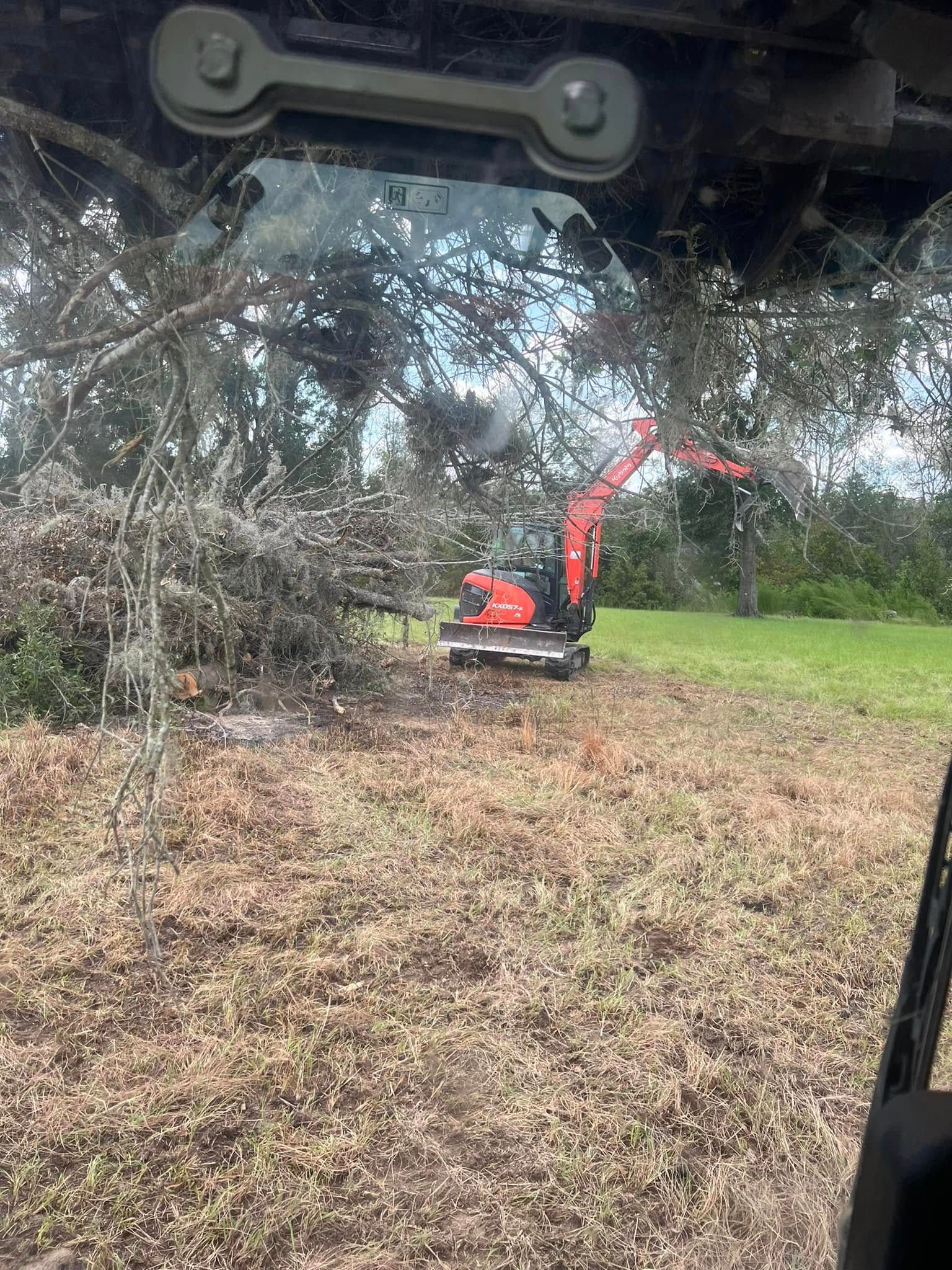 A bright orange excavator parked on a grassy field near a large pile of tree branches, viewed through a cab window.