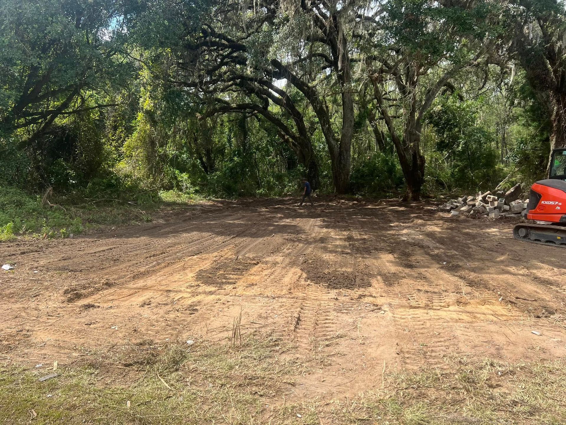 A leveled dirt patch in a wooded area with a small red excavator parked on the right.
