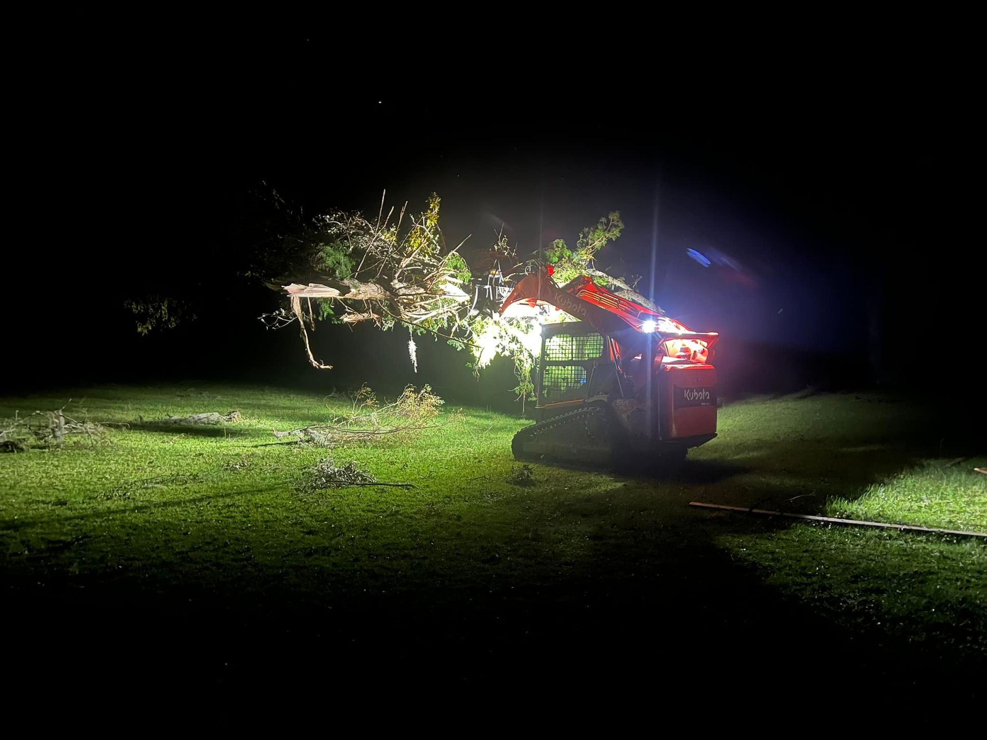 A red skid steer loader illuminates and clears fallen tree branches on a grassy field at night.