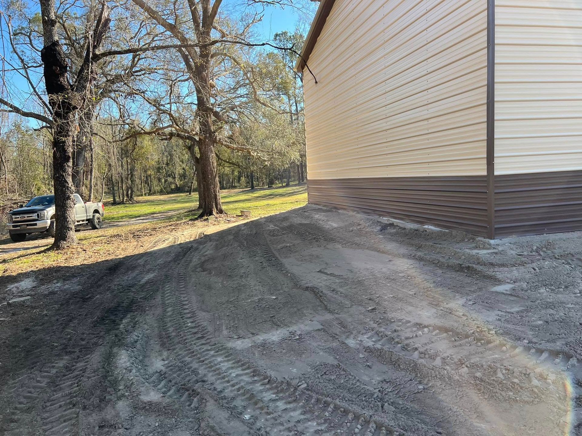 A corner of a beige building with brown trim sits next to a dirt driveway with tire tracks, near trees in a rural setting.