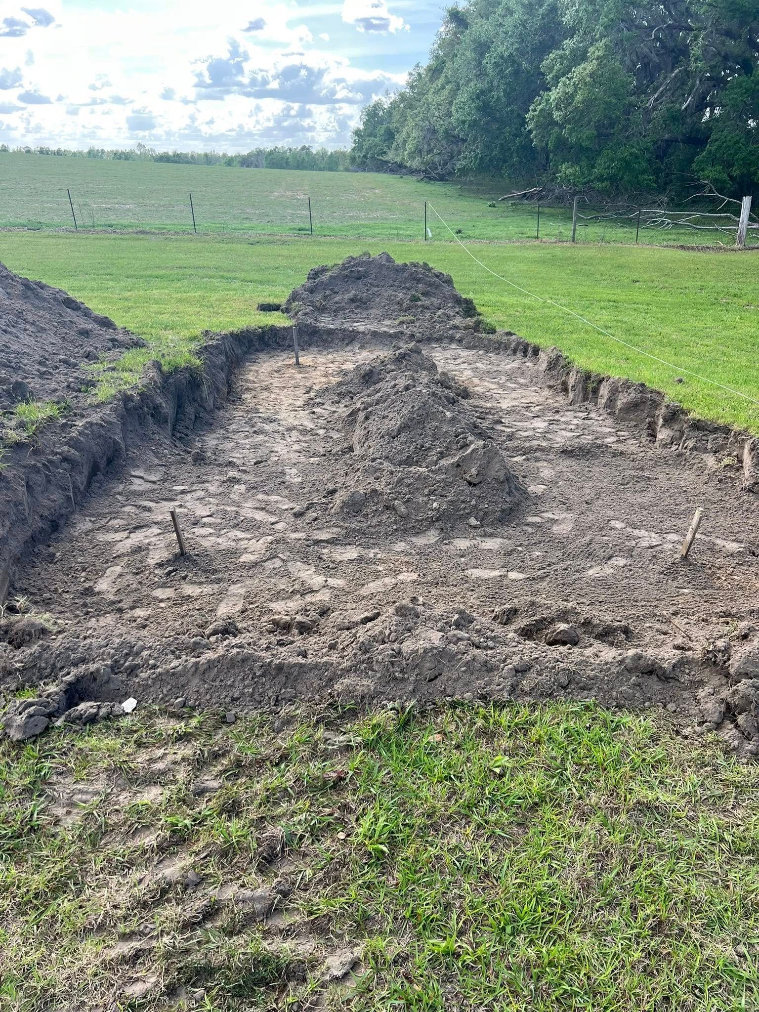 A partially excavated patch of dark earth in a grassy field under a cloudy, bright sky.