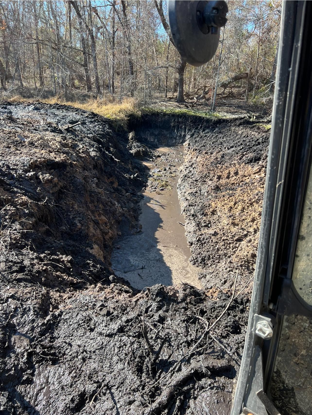A view from a construction vehicle looking down into a deep, muddy trench excavated through a wooded area.