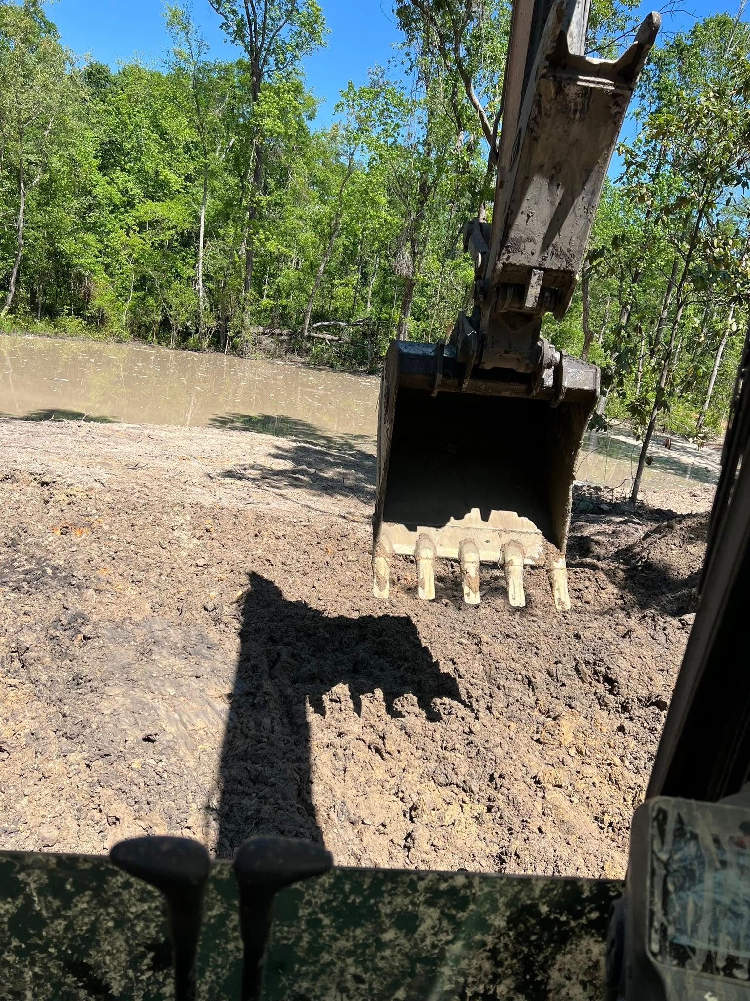 View from inside an excavator cab looking out at a large bucket suspended over a dirt field near trees under a blue sky.