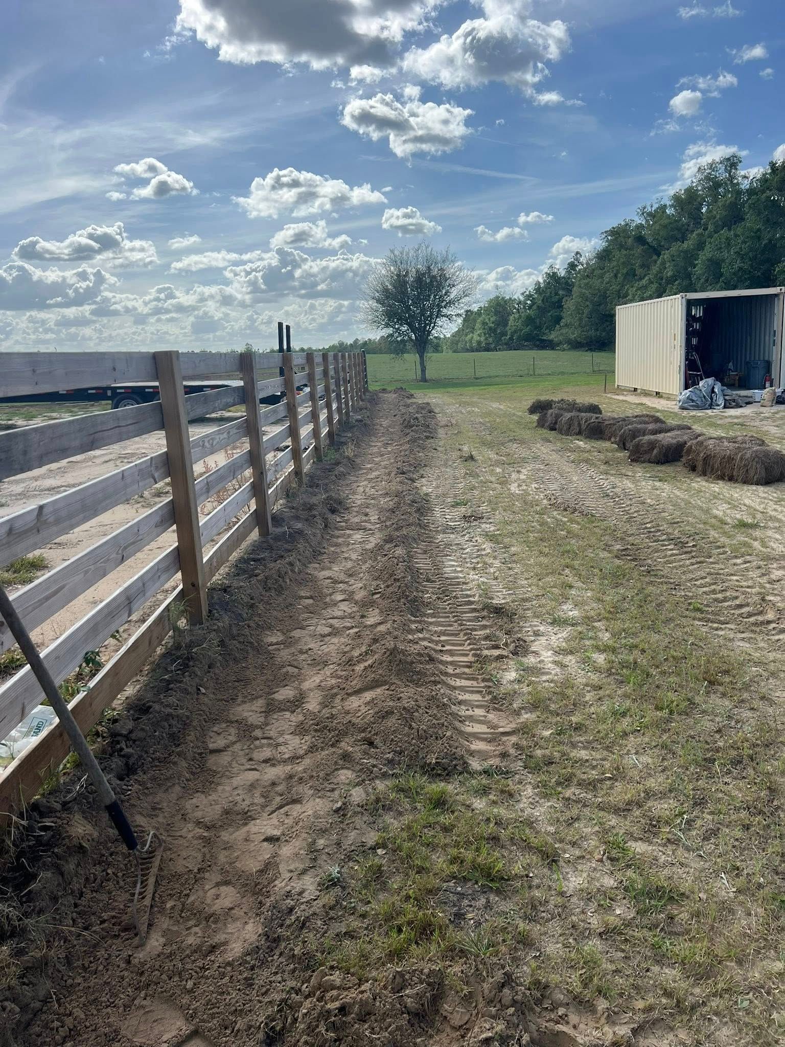A dirt trench runs alongside a long wooden fence in a rural field under a blue, cloudy sky.