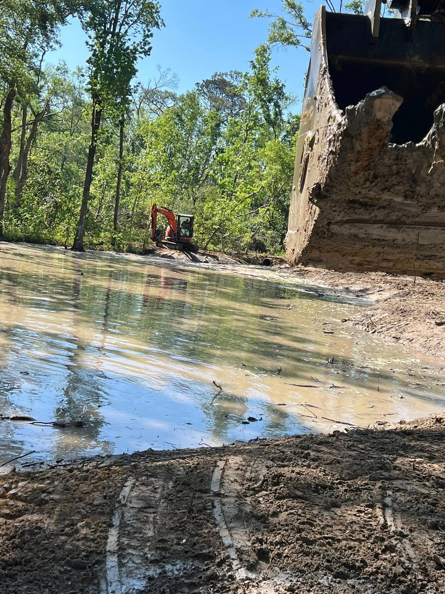 A large excavator bucket hangs in the foreground over a muddy, water-filled construction site with a distant excavator.
