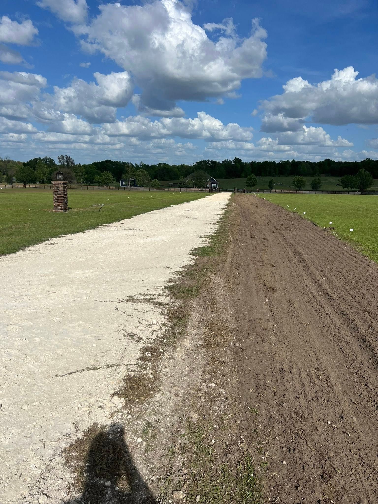 A gravel path runs beside a strip of freshly tilled dark soil in a green field under a blue sky with white clouds.