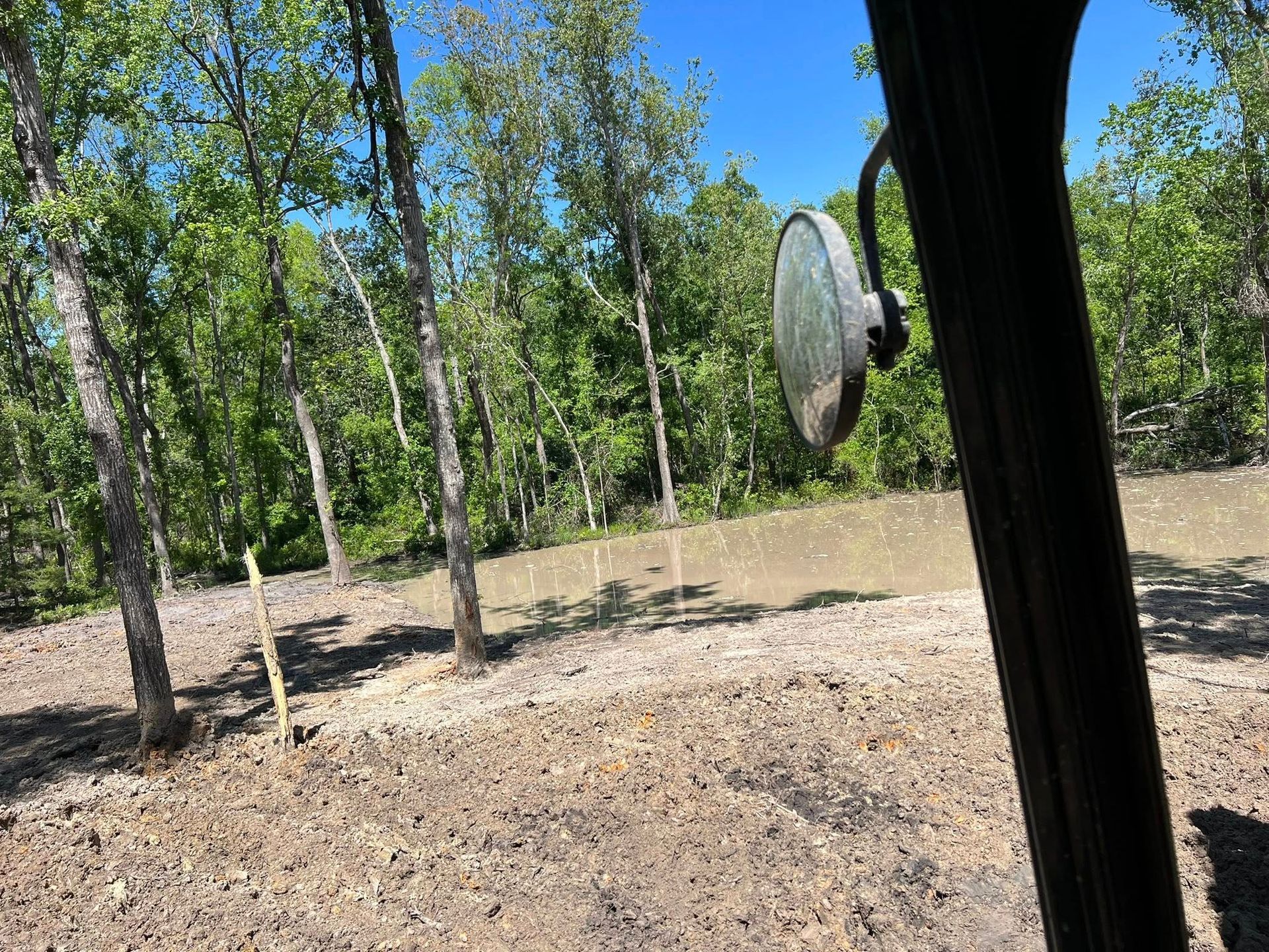 A side mirror view from a vehicle showing a muddy pond surrounded by green trees on a sunny day.