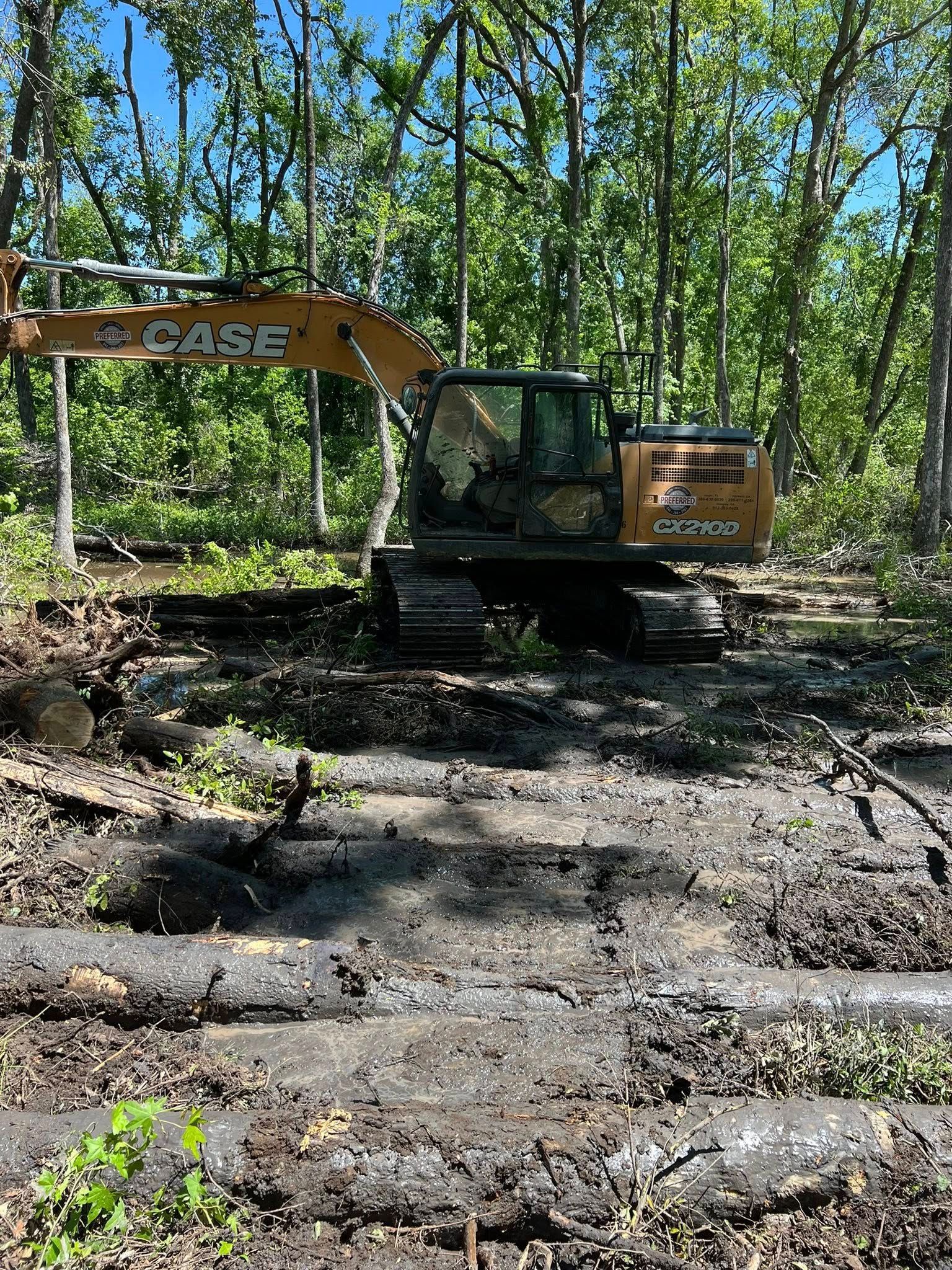 A yellow Case excavator sits in a muddy, wooded clearing surrounded by trees and debris.