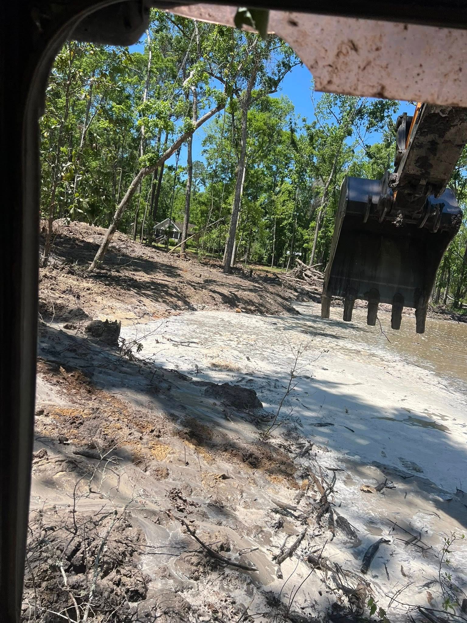 An excavator bucket looms over a muddy, cleared slope at the edge of a forest under a bright blue sky.