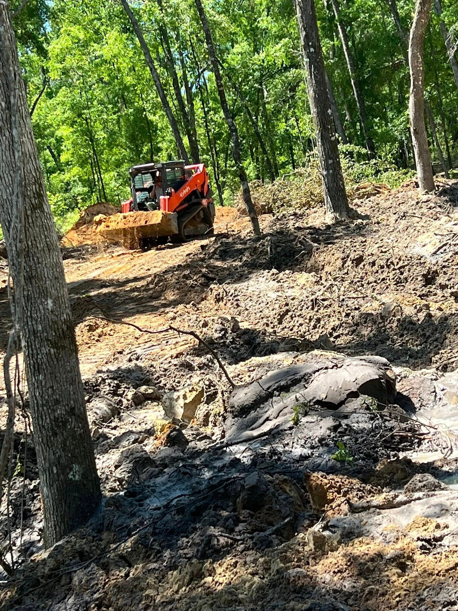 An orange skid steer loader clears dirt and vegetation in a wooded area on a sunny day.