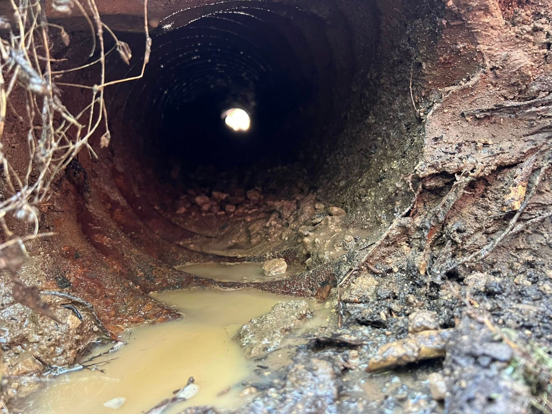 A dark, corrugated culvert pipe partially filled with muddy water and debris, with a bright light visible at the end.