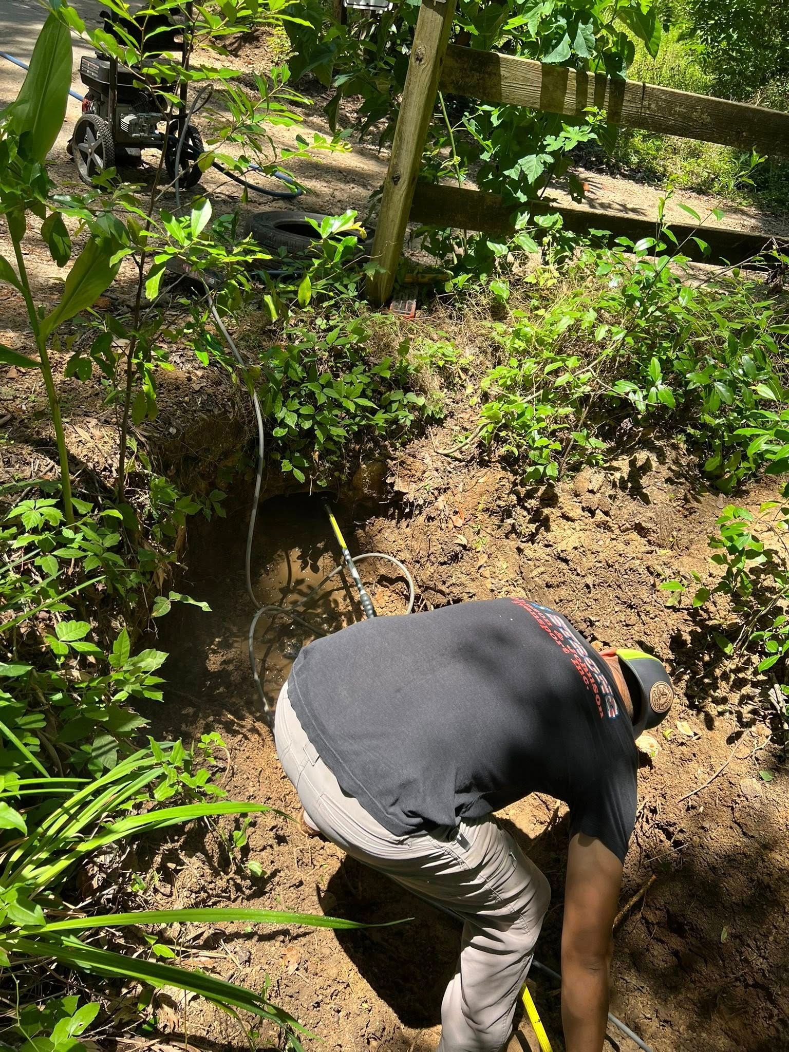 A person in a gray shirt and light pants works in a wooded area, digging near a small, muddy pool of water.