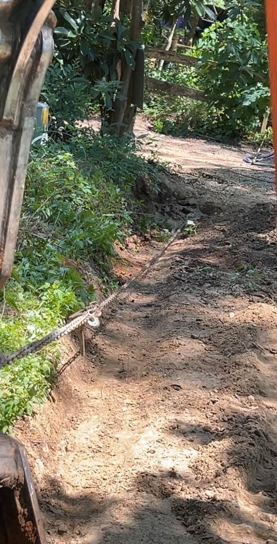 An orange excavator arm overlooks a narrow dirt trench being dug along a path with greenery and trees in the background.