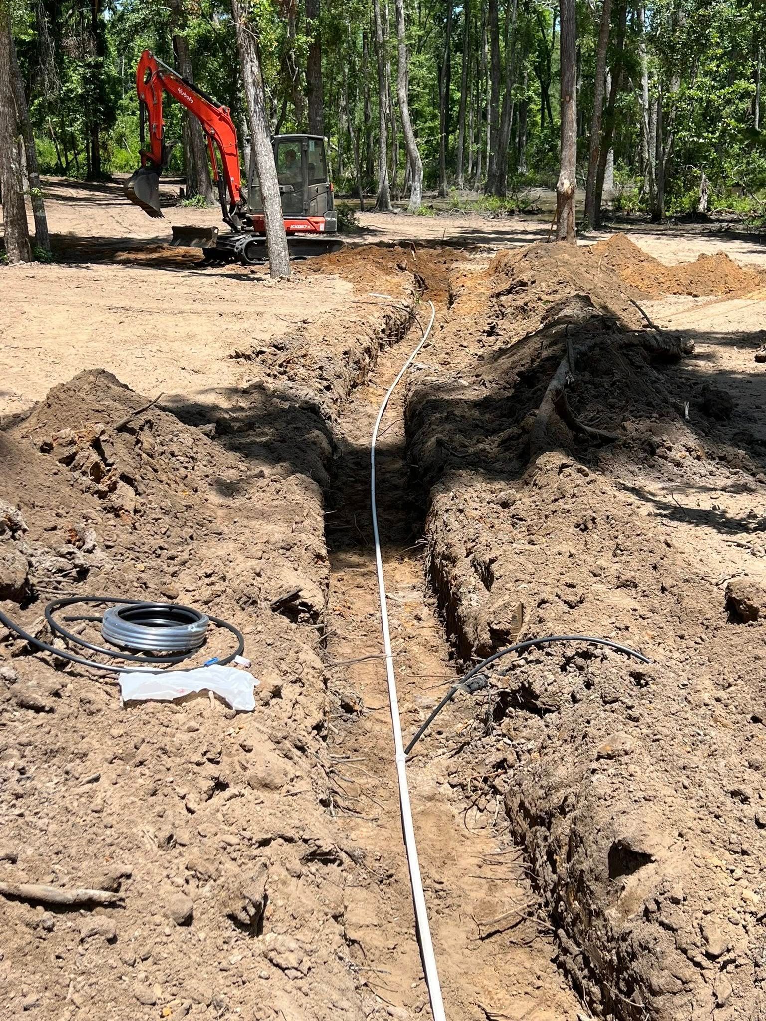 An orange excavator sits in a wooded area behind a freshly dug trench with a white pipe laid inside.