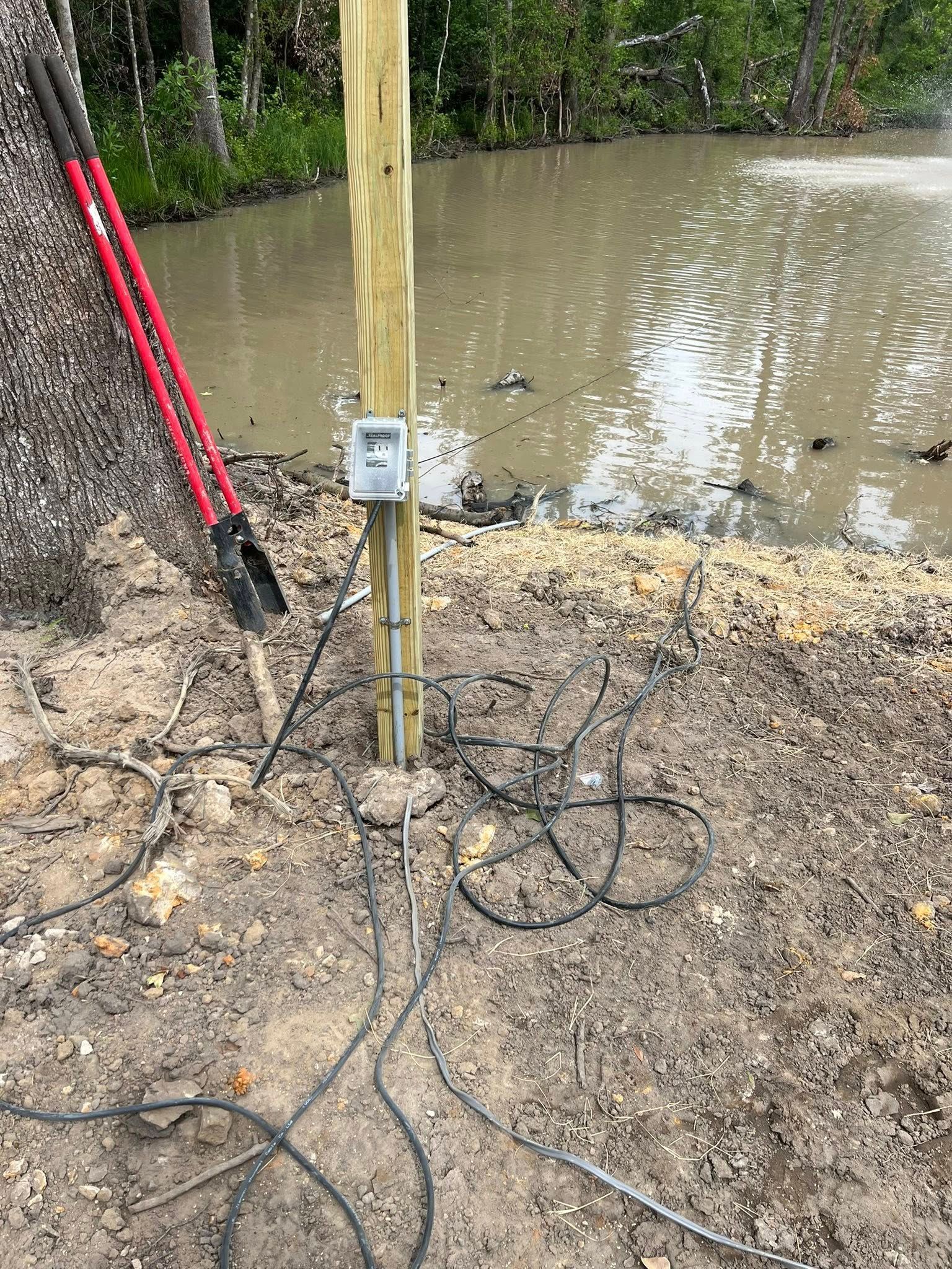 A red-handled shovel leans against a wooden post with an electrical box near a pond edge, surrounded by loose cables.