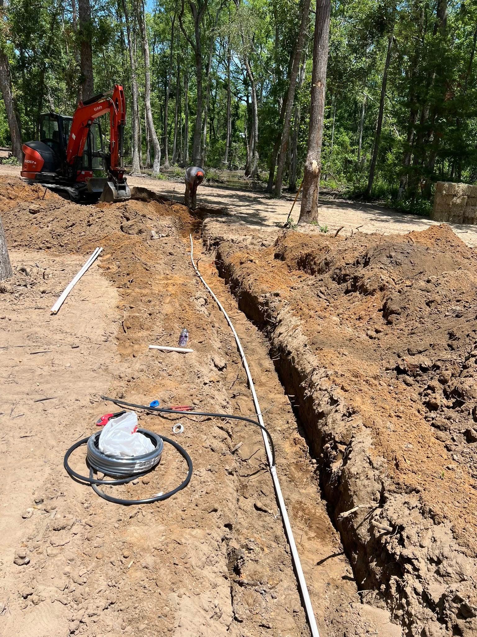 A red excavator sits near a long, open trench in a dirt field surrounded by trees, with a coiled pipe in the foreground.