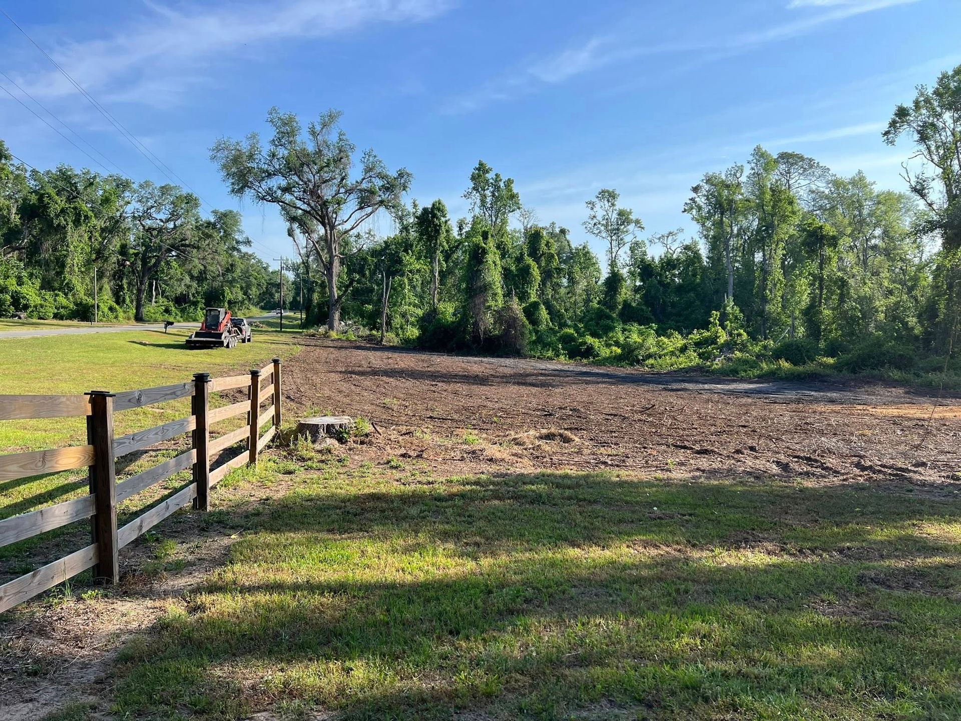A wooden fence lines a field where a tractor works to clear land near a line of trees under a bright blue sky.