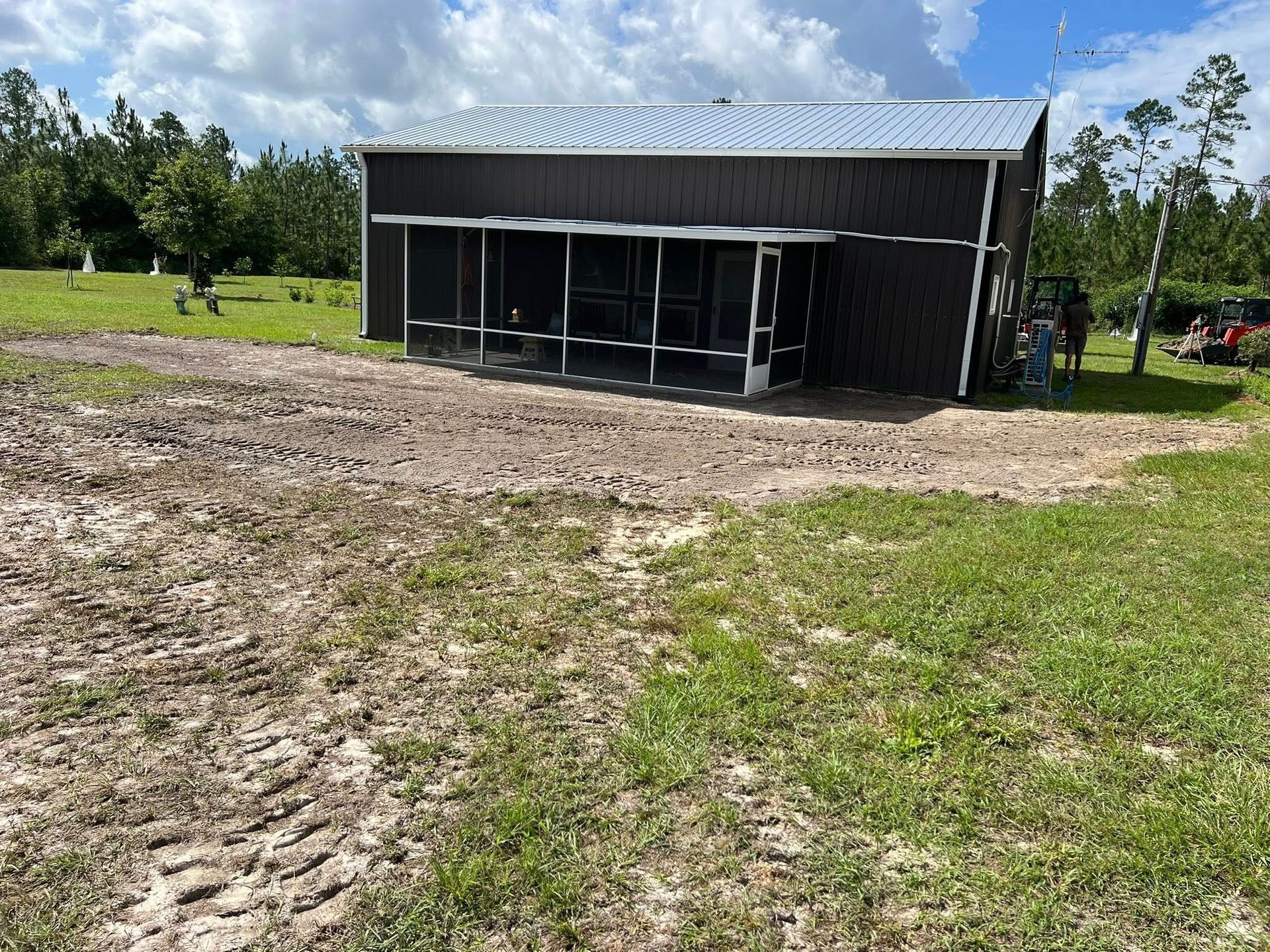 A dark metal building with a screened porch sits in a large, rural grassy field with disturbed soil in the foreground.
