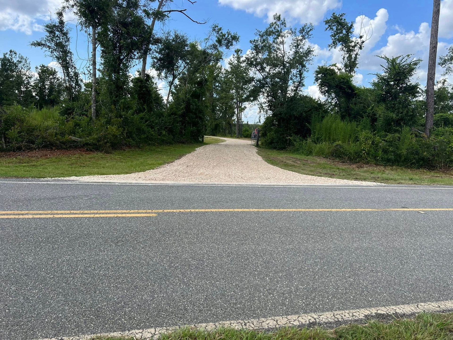 A paved road with a yellow double line meets a light-colored gravel driveway that leads into a wooded area.