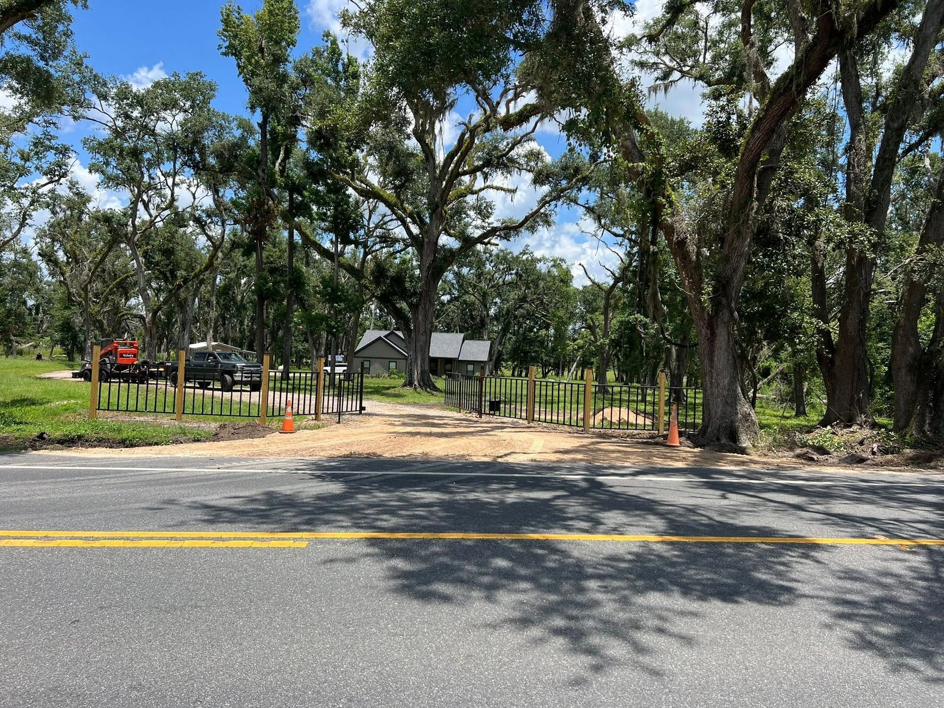 A gravel driveway leads from a paved road into a wooded area with several tall, thin posts marking the entrance.