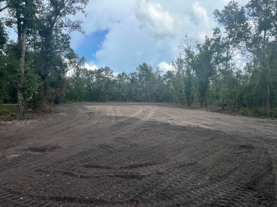 A clearing in a forest with freshly turned dark soil and tire tracks leading toward a patch of light-colored gravel.