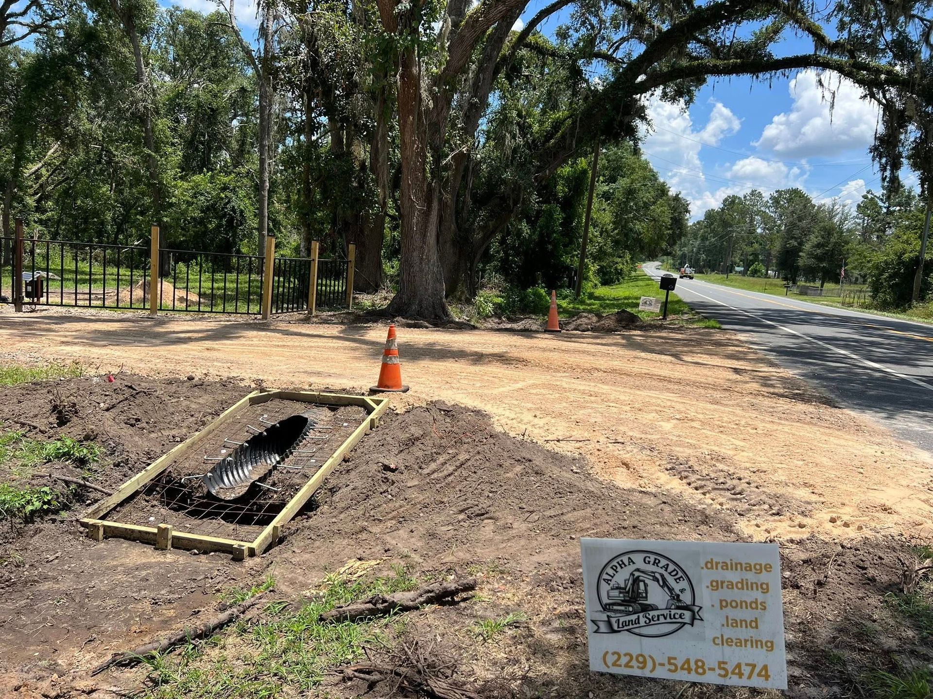 A roadside construction site with a wooden-framed drainage pipe, a traffic cone, and a contractor sign on sandy ground.