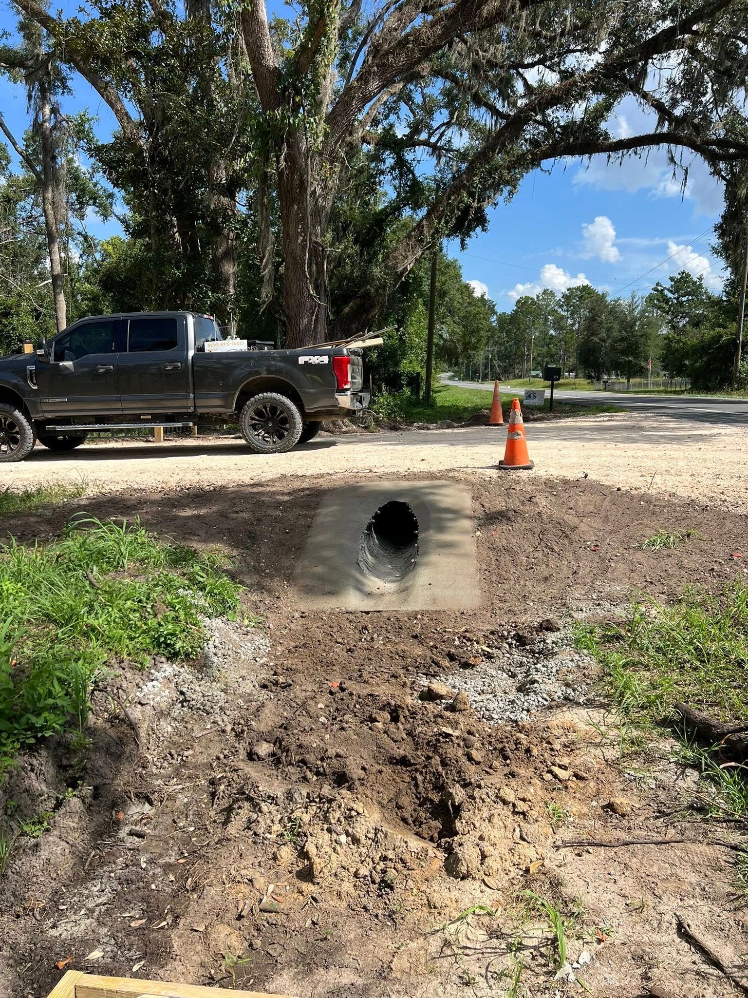 A drainage pipe set in a concrete base beside a gravel road, with a dark pickup truck parked nearby under trees.