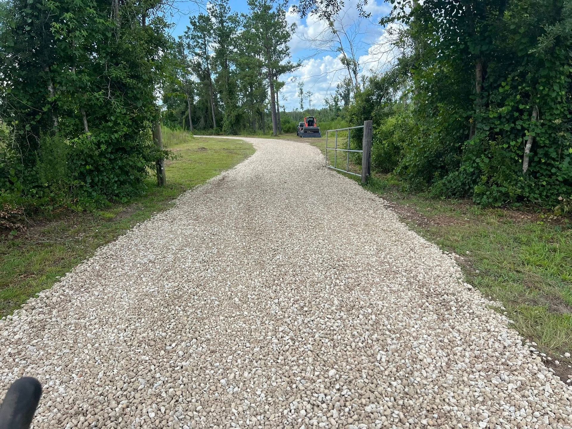 A gravel path winding through a grassy, tree-lined landscape under a bright, partly cloudy sky.