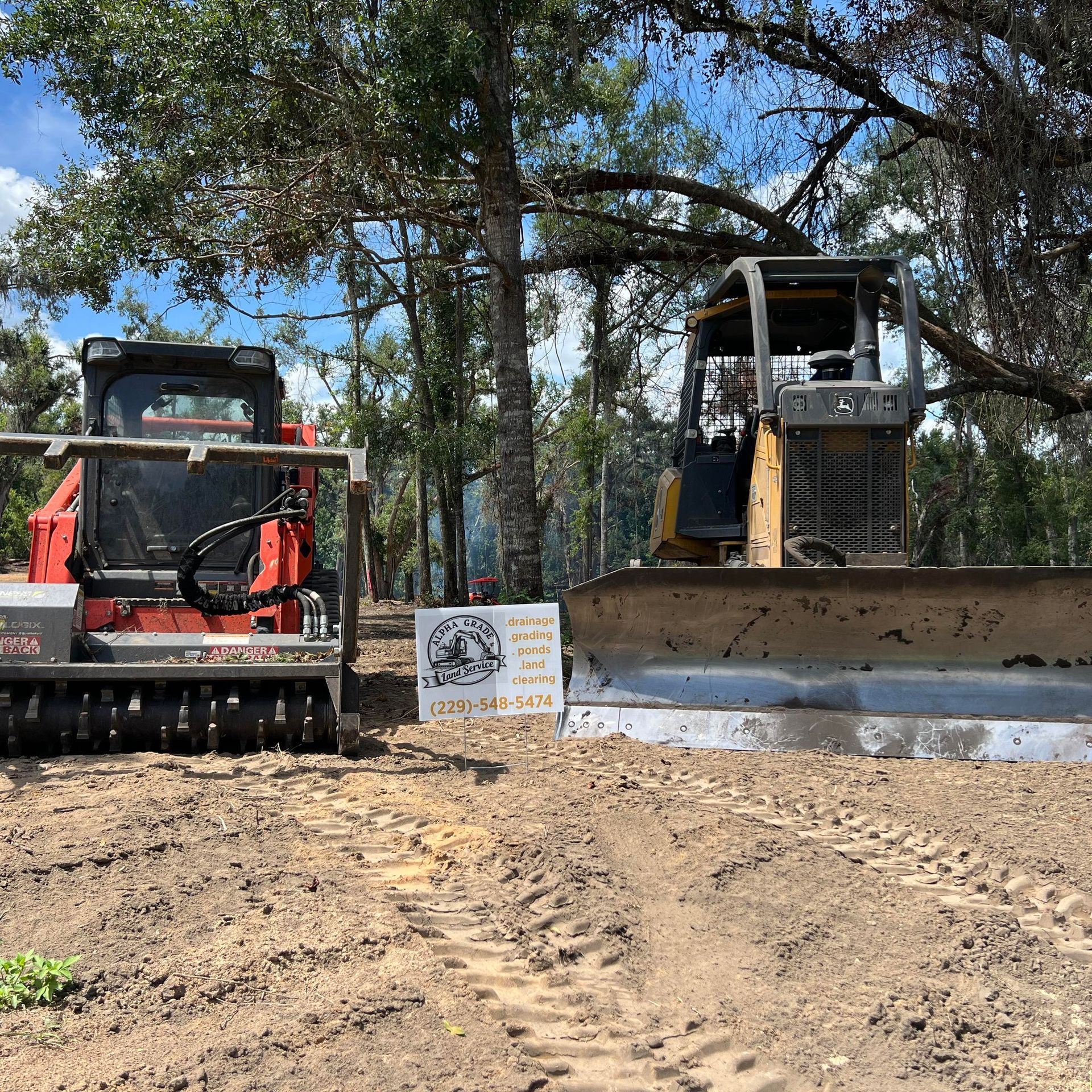 A red forestry mulcher and a yellow bulldozer parked on dirt ground with a sign between them under trees.