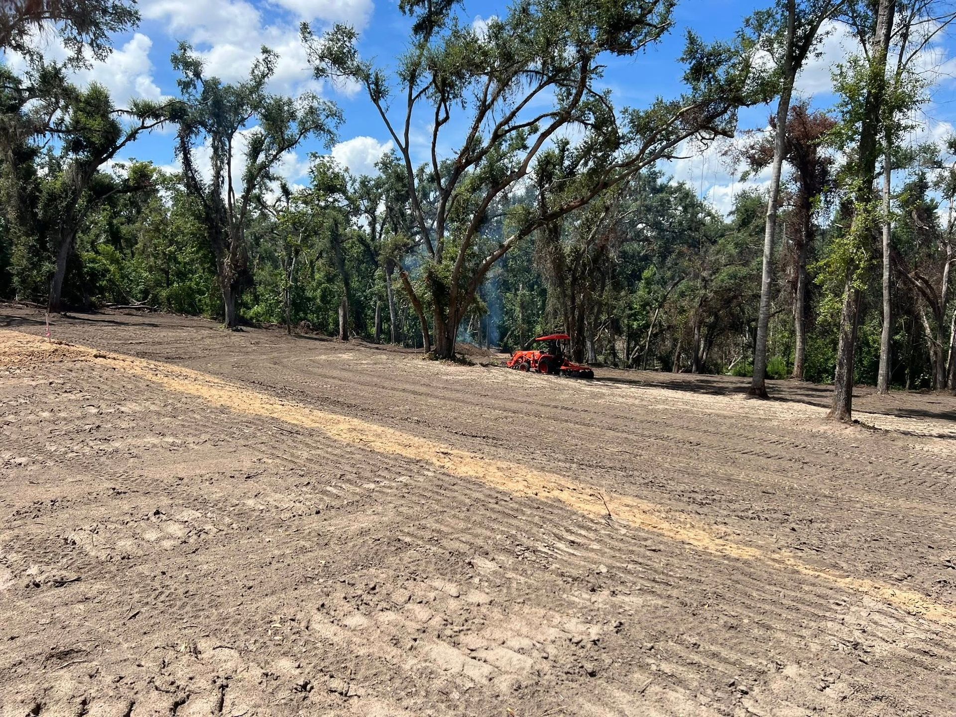A small orange tractor sits on a cleared, dirt-covered lot near a line of trees under a blue, partly cloudy sky.