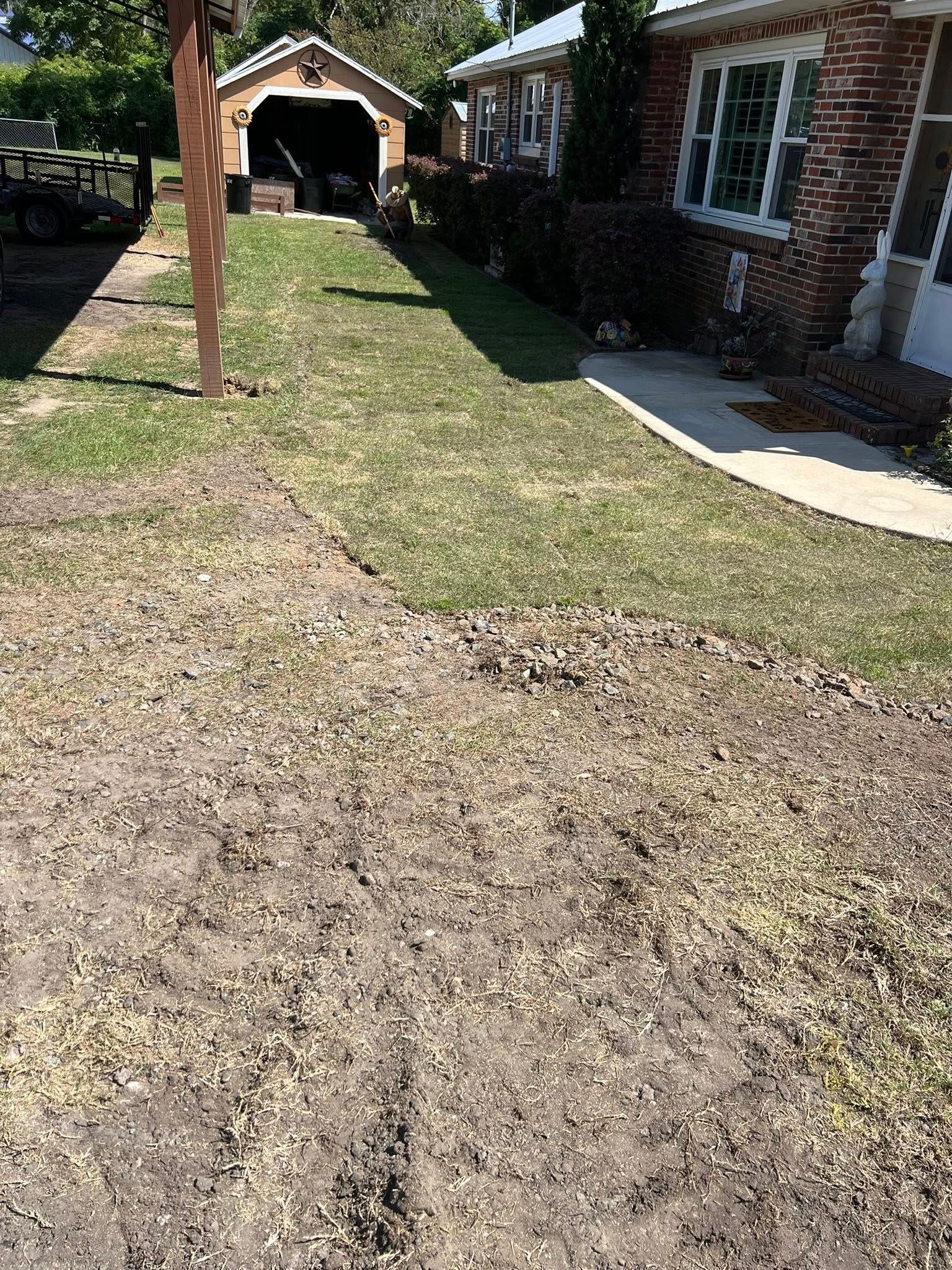 A partially shaded yard transitioning from bare dirt in the foreground to green grass leading to a brick home and garage.