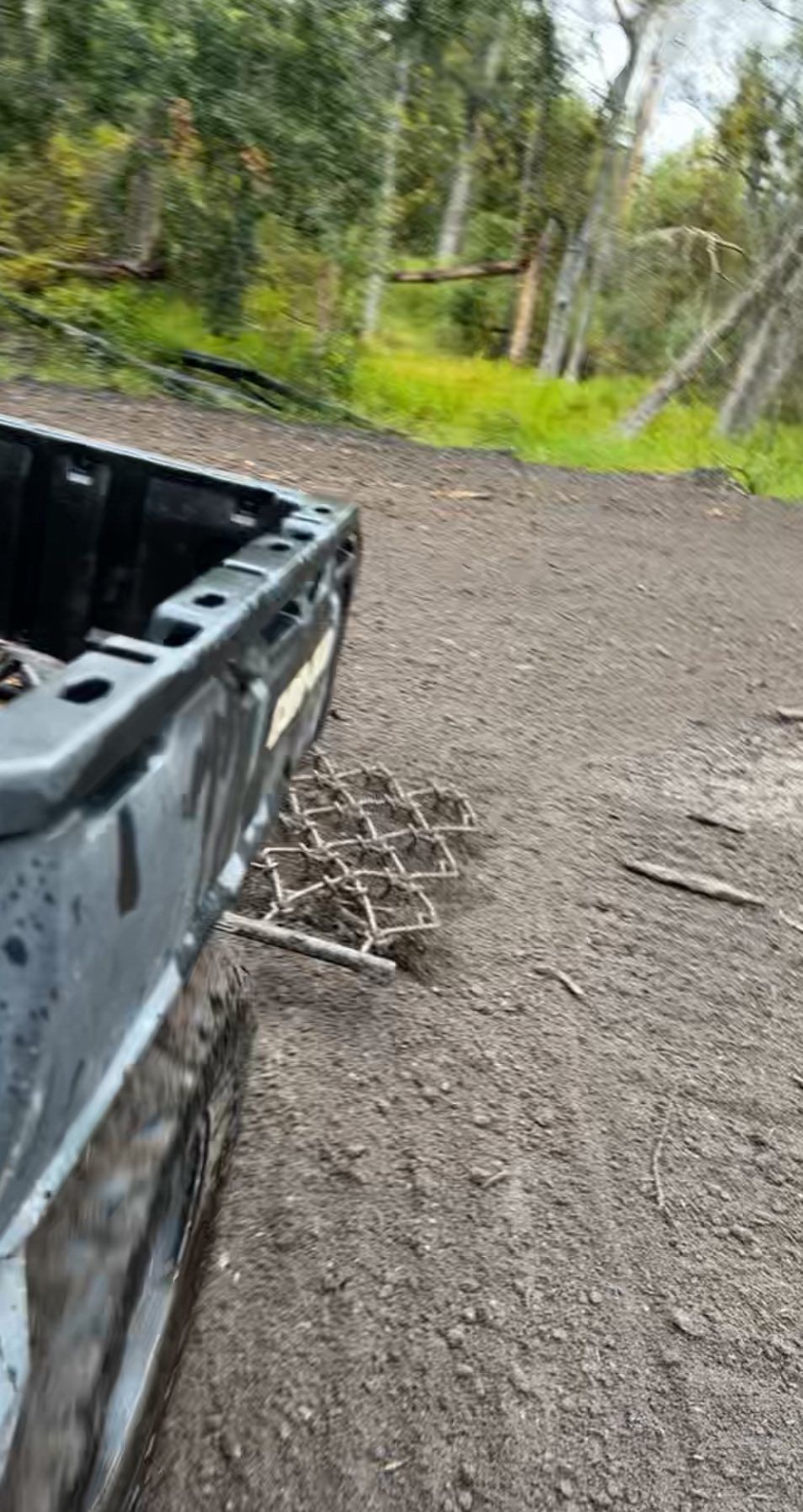 A metallic drag harrow attached to the back of an ATV, parked on a dirt path near a wooded area.