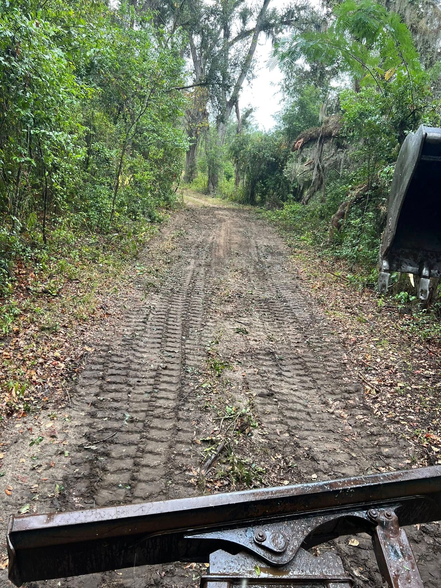 A muddy forest trail marked by deep, wide, parallel tire tracks, viewed from the perspective of a vehicle's front blade.