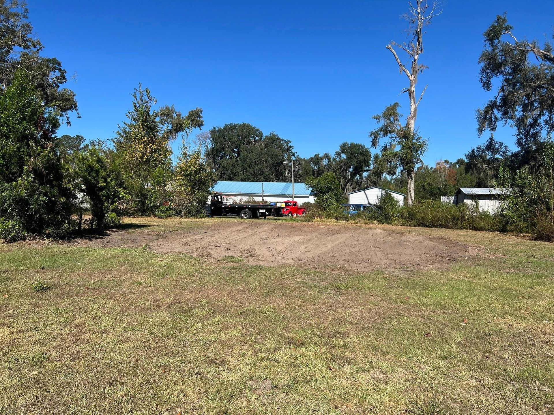 A patch of cleared, bare dirt in a grassy field, with trees and a parked truck in the background under a blue sky.