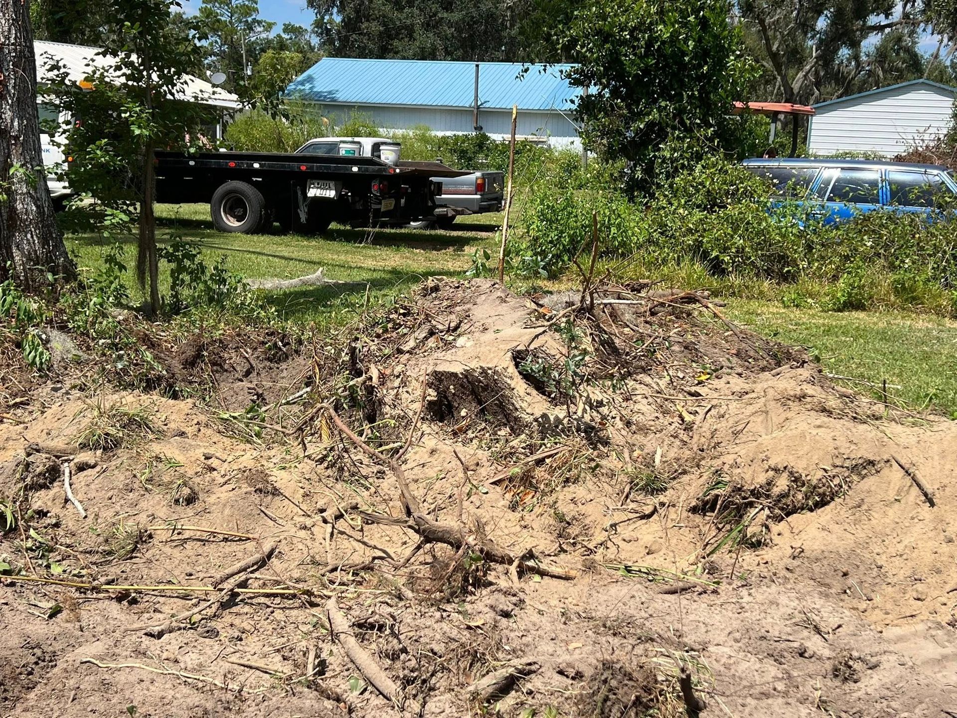 A dirt lot with debris and a large stump, featuring a black trailer and blue building in the background.