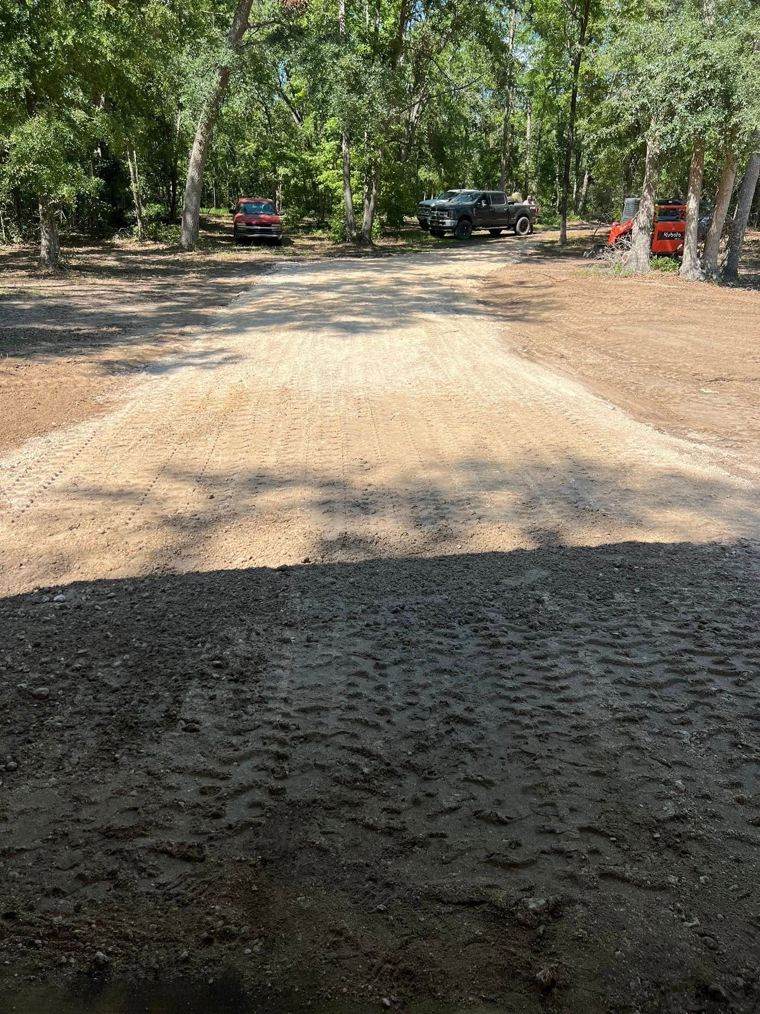 A gravel driveway stretches into a wooded area where several parked vehicles are visible in the background.