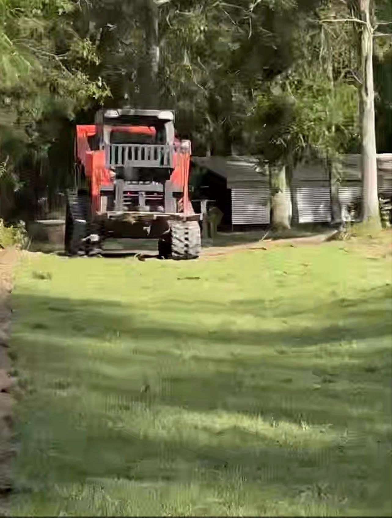 An orange tracked skid steer loader parked in a grassy field in front of a small shed surrounded by trees.