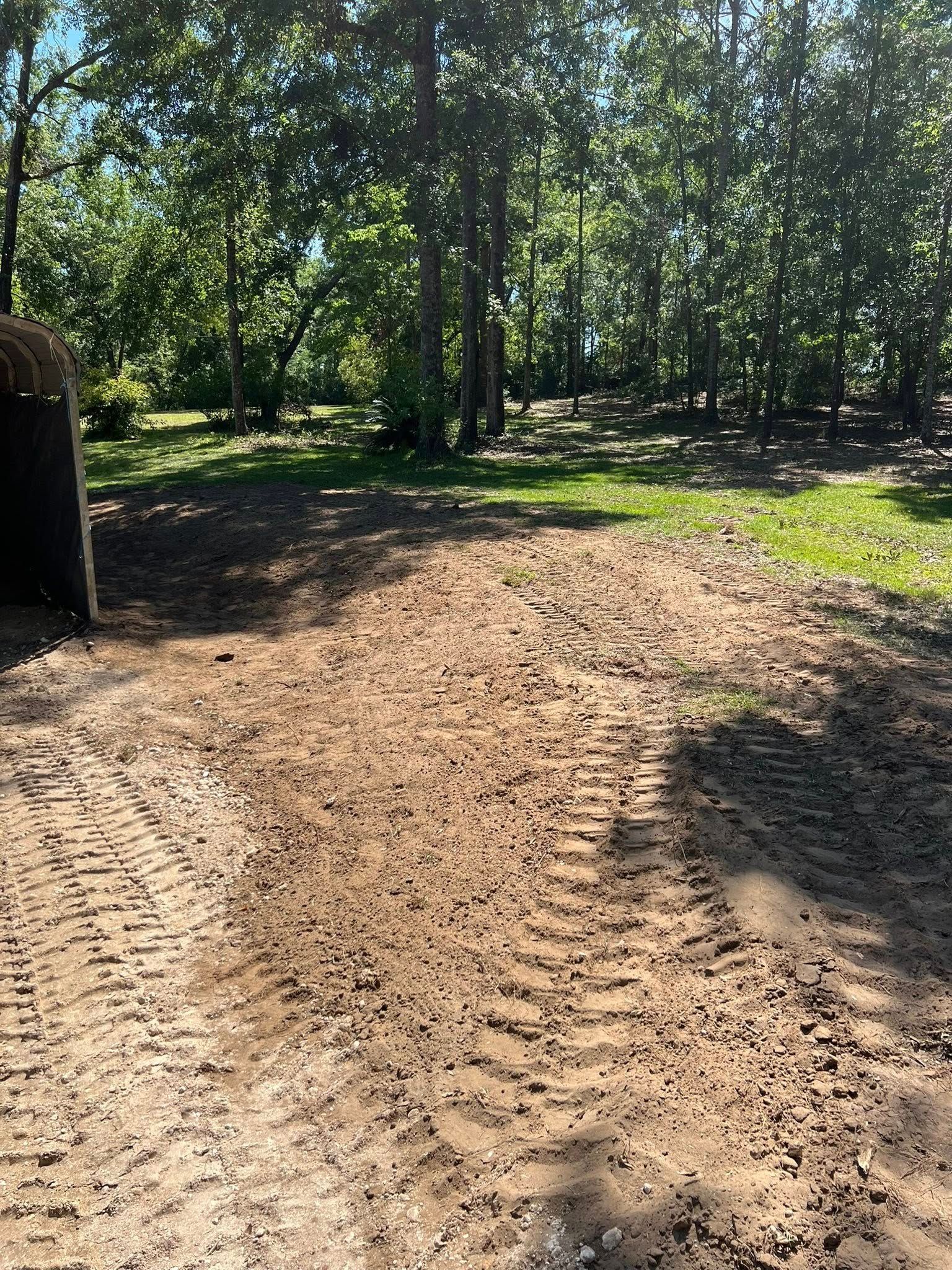 Freshly tilled dirt ground with deep tire tracks, positioned in front of a tree-lined forest area on a sunny day.