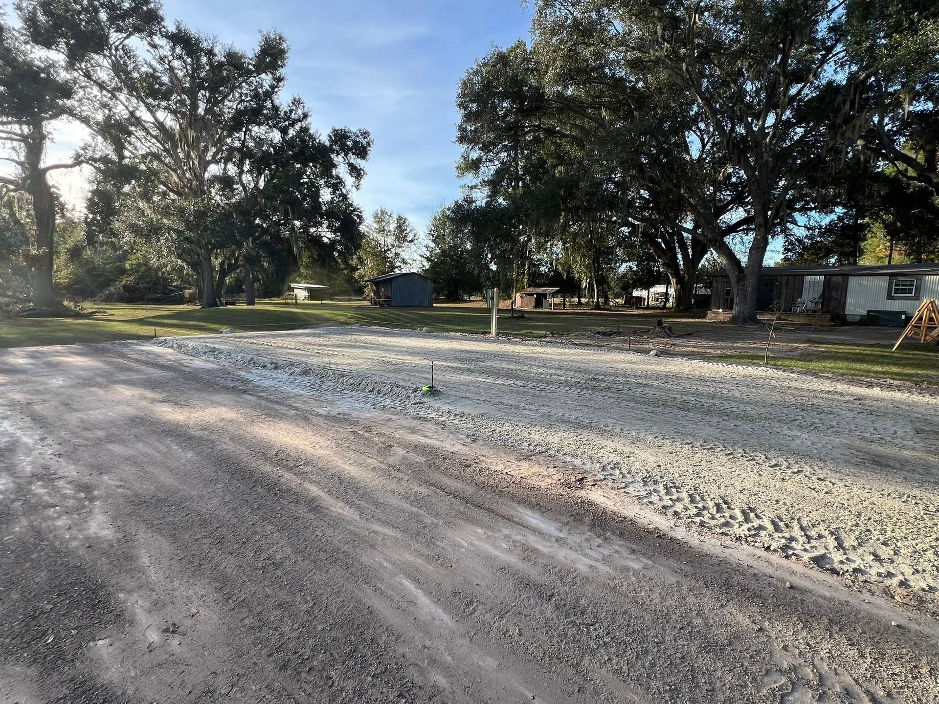 A gravel driveway leading toward a grassy yard with trees and several structures in the distance on a sunny day.