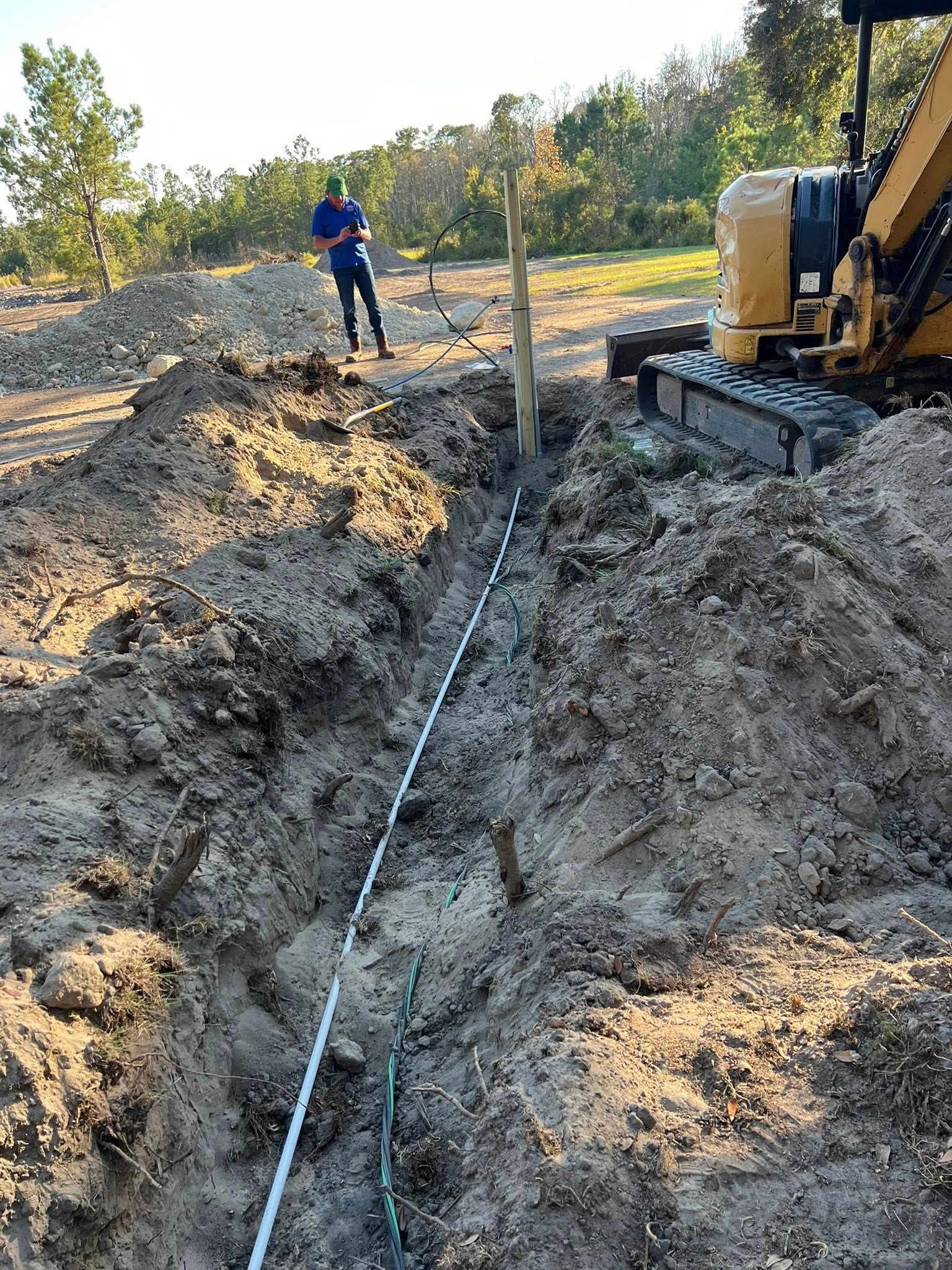 A person stands near a trench being dug by a construction machine, with a line of cable laid along the ground.