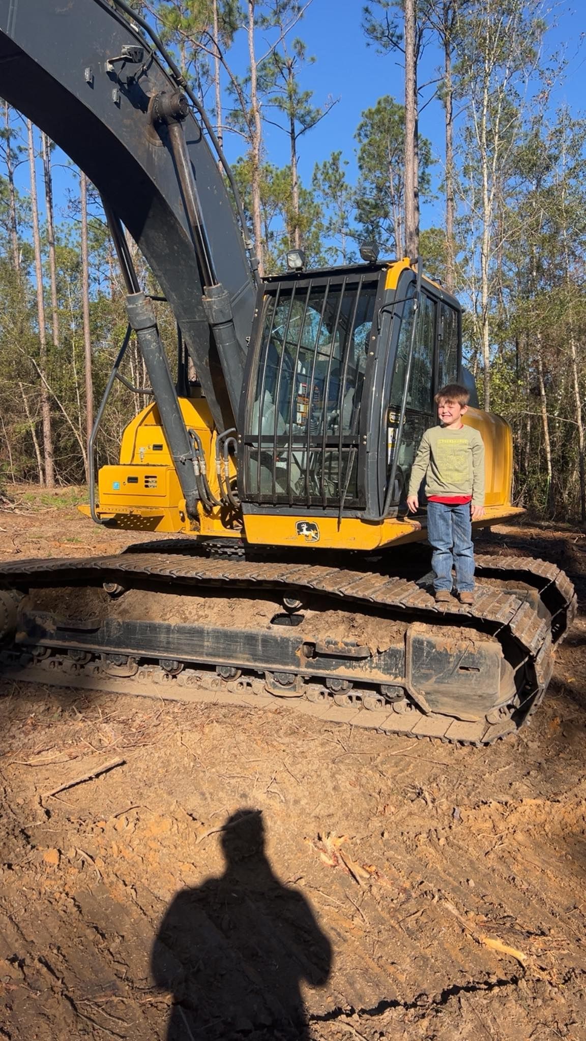 A young child stands on the yellow metal tracks of a large, dark-colored forestry excavator in a wooded area.