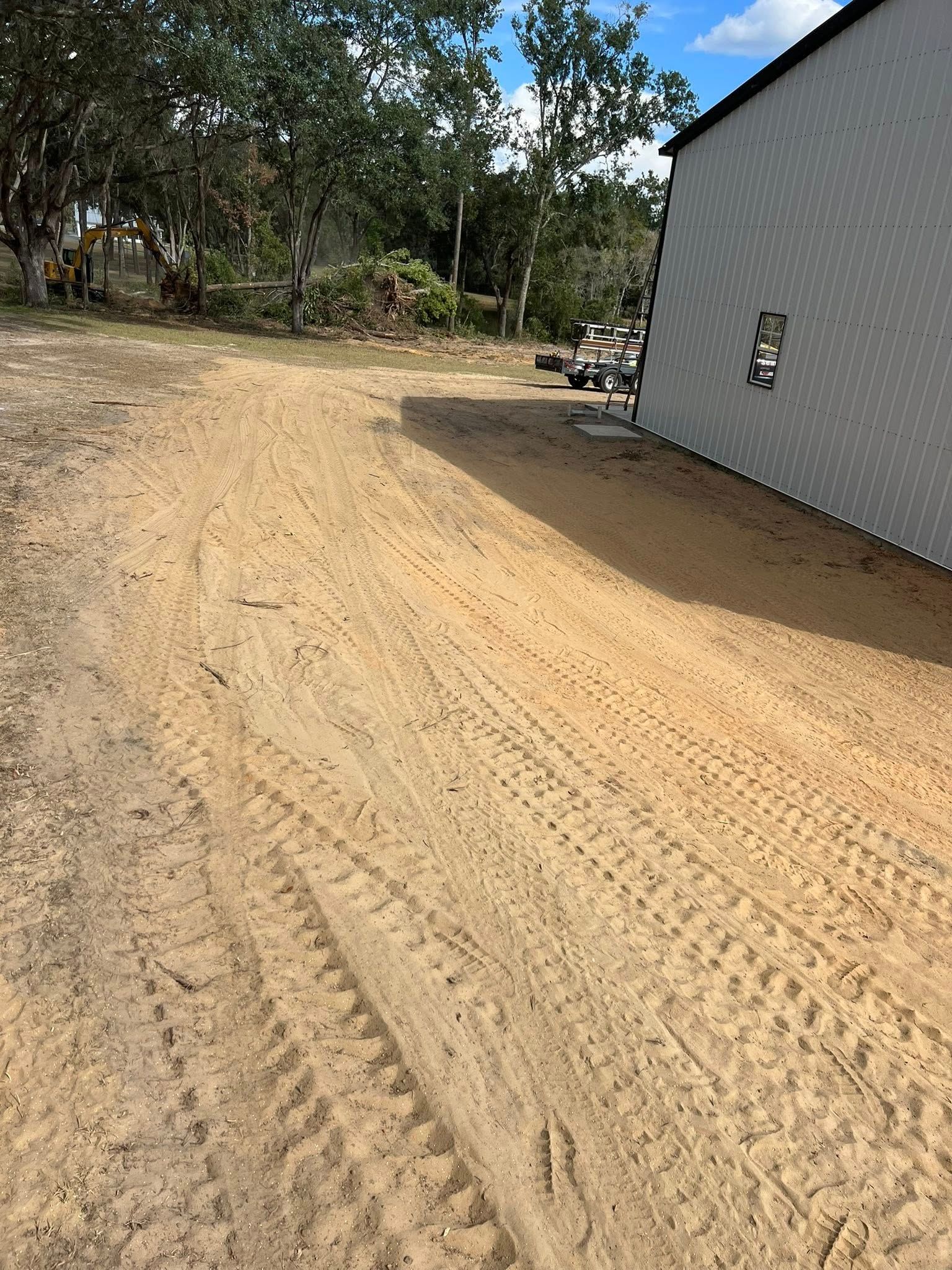 A sandy dirt path curves around the side of a white metal building, with trees in the background under a blue sky.
