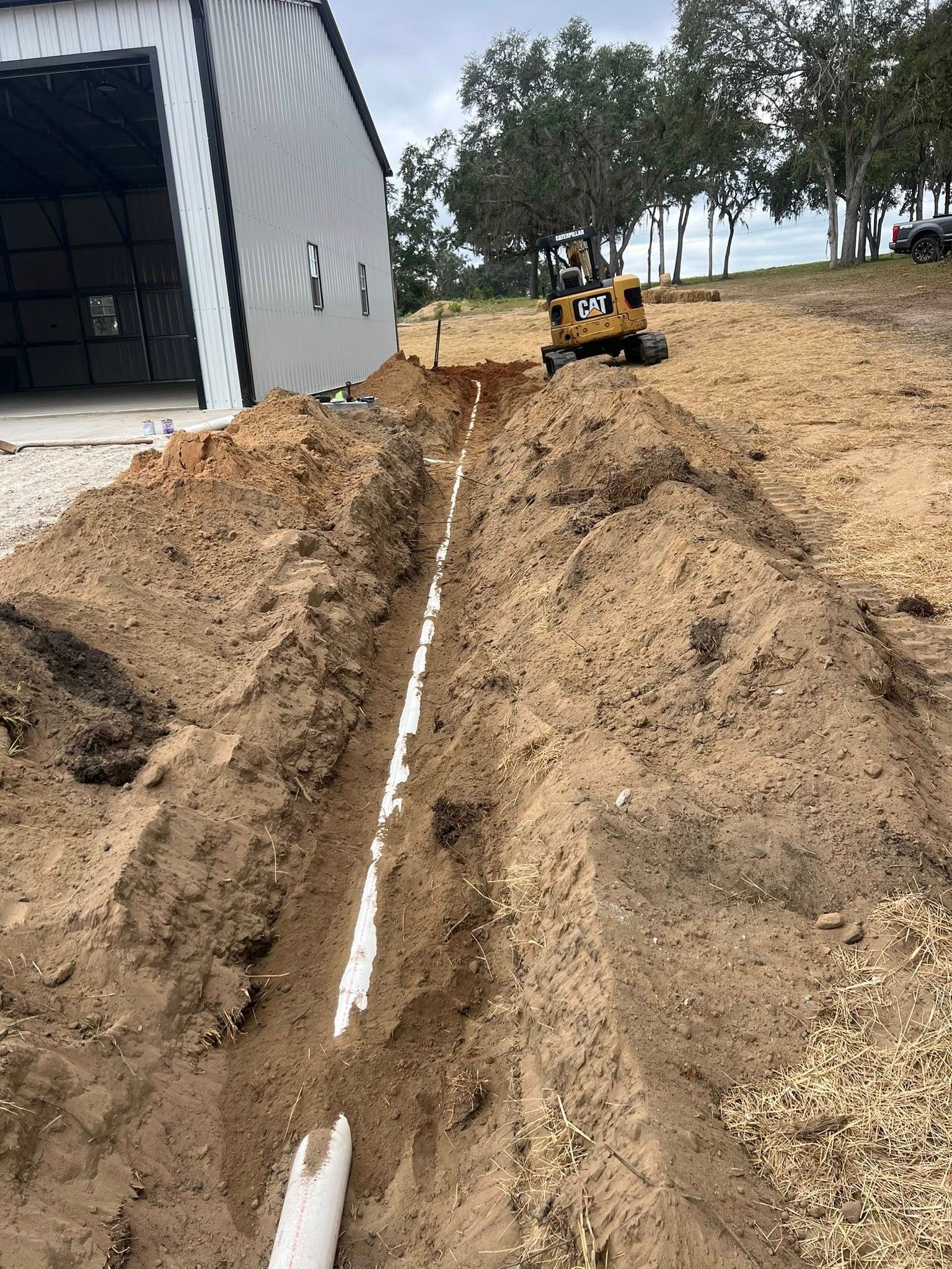 A yellow Caterpillar excavator sits at the end of a long, deep trench containing a white drainage pipe beside a metal shed.