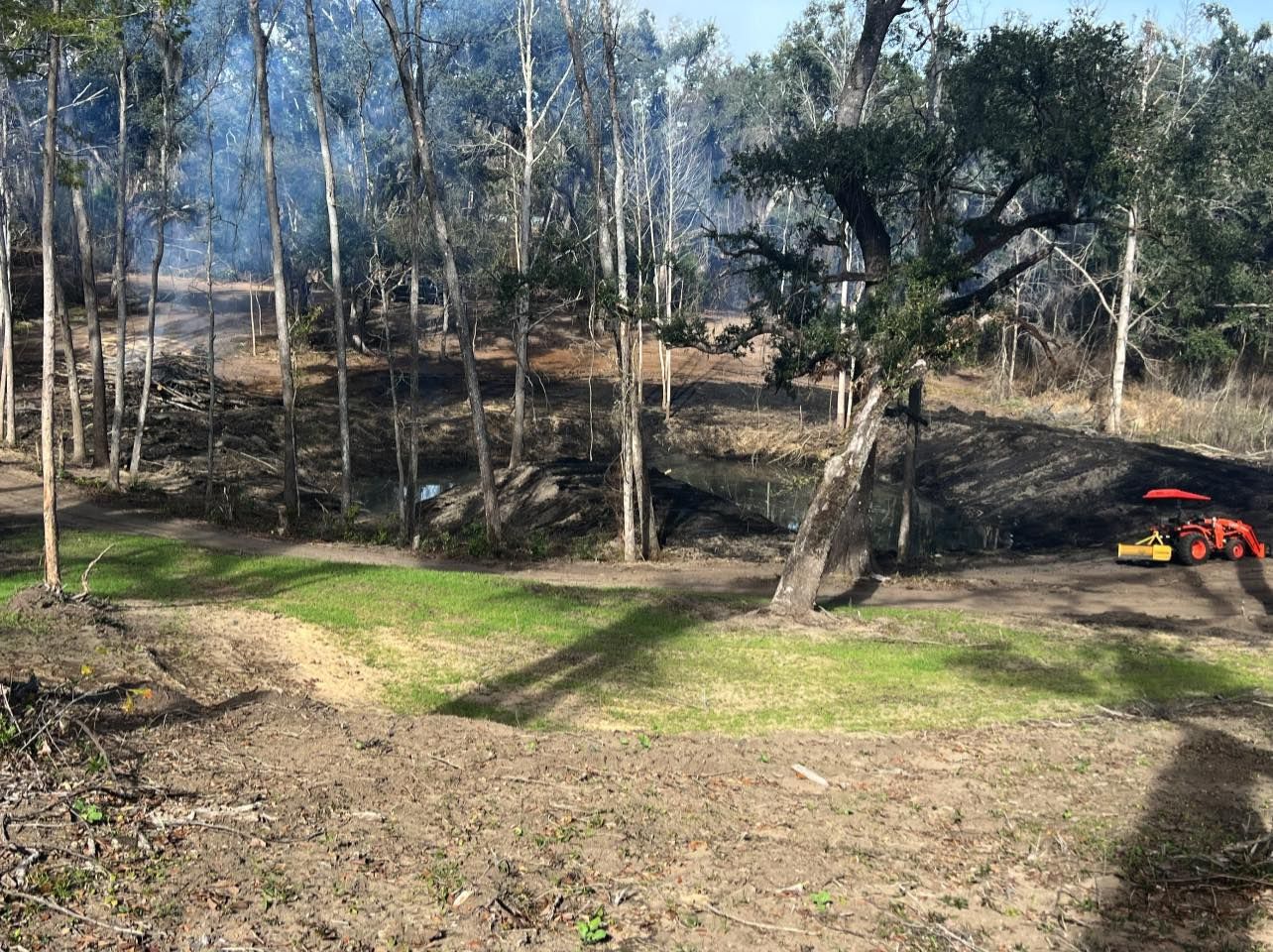 A bright orange tractor works in a wooded area with patches of green grass, smoky ground, and dark, scorched earth.