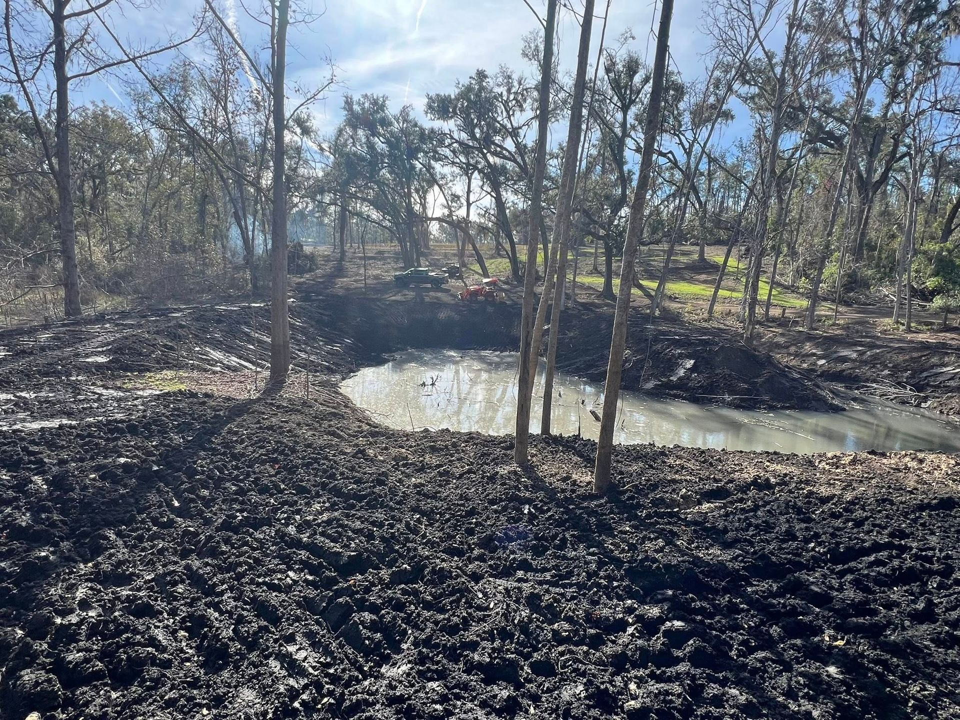 A muddy, churned-up construction site surrounding a small, murky pond in a wooded area under a bright sky.