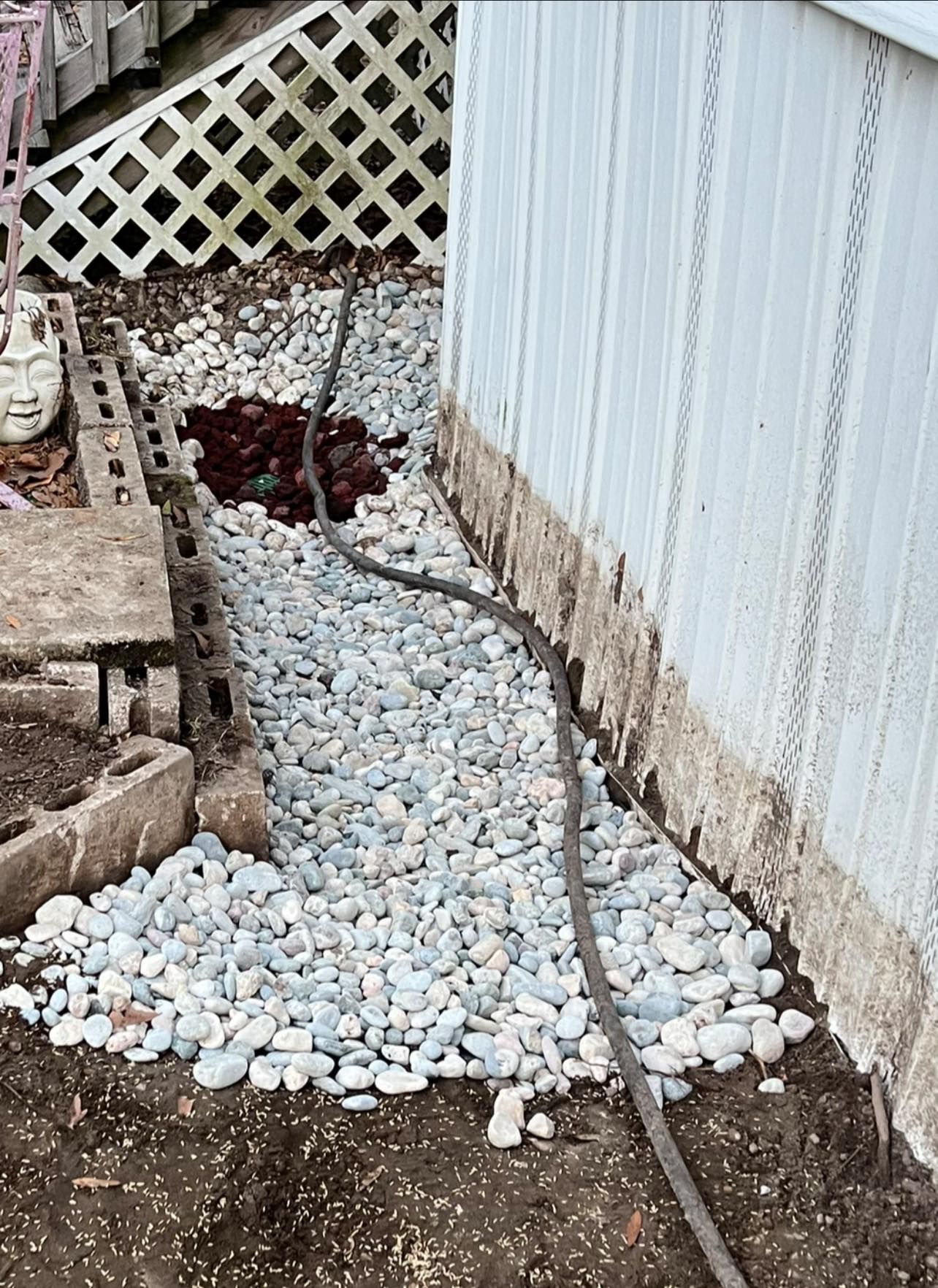 A garden bed alongside a white building wall features a path of light-colored river stones and a dark water hose.